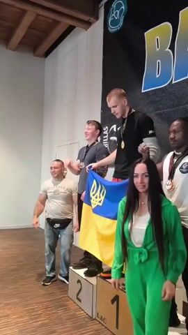 A group of five people are standing on a podium. A Ukrainian flag is draped over the shoulders of the person standing on the first-place platform. The person on the first-place platform is a young white male wearing a black t-shirt, gold medal, and orange pants. To his left, on the second-place platform, is a young white male wearing a dark t-shirt and a gold medal. To the right of the first-place person, on a lower platform, is a young Black male wearing a white t-shirt, a gold medal, and black pants. To the left of the second-place person, on the third-place platform, is a white male wearing a light-colored t-shirt and jeans. A young white female in a green tracksuit is standing to the right of the first-place person, slightly in front of the podium. The background is a black banner with a logo and the word "BABY" in large letters. The podium has numbers 1, 2, and 3 on the platforms. The environment appears to be indoors, possibly a sports venue.