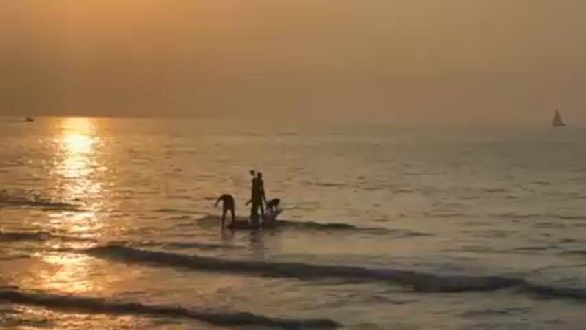 The scene is a wide shot of a calm ocean at sunset. The sun is setting on the left, casting a golden glow across the water. Two people stand on a paddleboard closer to the camera, and a dog is also on the board. One person holds a paddle. Gentle waves roll toward the viewer. In the distance, a sailboat is visible on the right, and another small boat is on the far left. The overall tone is warm and serene.