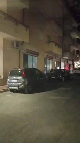 The video captures a street scene at night. Several cars are parked along the side of a building. A person wearing a white t-shirt and blue jeans is walking away from the camera on a sidewalk. The building appears to have shops or businesses on the ground floor, indicated by windows and signs. Streetlights and lights from the building illuminate the area. The camera movement is shaky, suggesting it might be handheld.