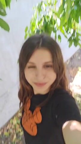 The video shows a young woman with long brown hair, winking and smiling at the camera. She is wearing a black t-shirt with an orange spiderweb design on it. The background is an outdoor setting with green leaves and a light-colored wall. In some shots, a bicycle is visible in the background. The woman is seen posing and interacting with the camera, at one point making a kissing face.