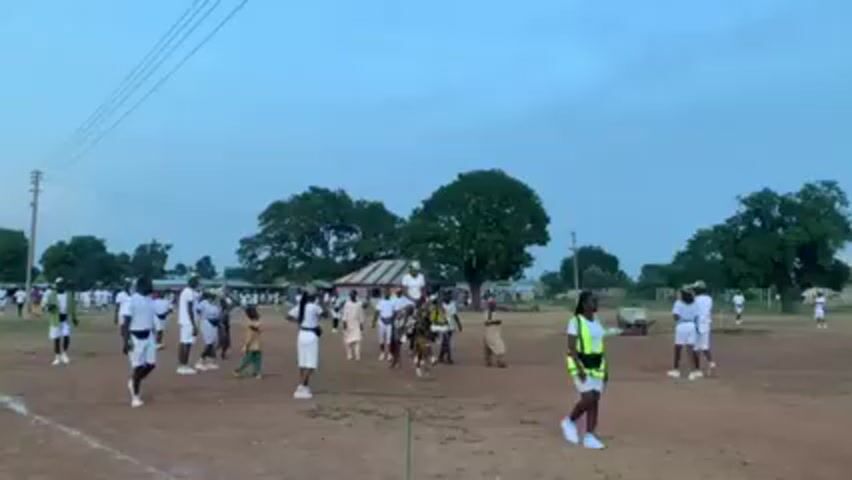 The video depicts a grassy field with several people gathered. Most are wearing white tops and bottoms. A person in white is riding a decorated horse, being led by another person. A wheelbarrow is being pushed nearby. The sky is overcast, and there are trees and buildings in the background. Some people are walking around, while others are standing.