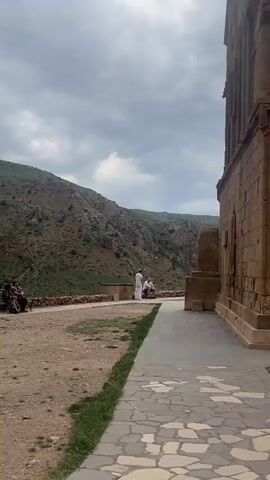 A low-angle, panning shot of people and a stone building on a dirt path with green hills and a cloudy sky in the background. A couple sits on a wooden bench at the start of the video. As the camera pans right, a woman in a maroon top and dark pants with a backpack walks past the camera. The video ends with a shot of the dirt path, the green hills, and a cloudy sky.