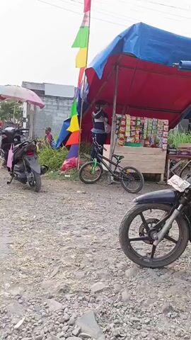 A roadside stall with a blue and red tarp canopy is set up. Various snacks are displayed on shelves. A person in a face mask stands behind the stall. A bicycle is leaning against the stall. Two motorcycles are parked nearby, one in the foreground and one behind the stall. A flag with colorful triangular panels flies from a bamboo pole in front of the stall. Further down the road, a woman in a black hijab hands a tray to a man in a teal shirt and cap, who is sitting on a low wall. Two other men are sitting on the ground under trees; one is looking at his phone, and the other is looking towards the camera. The environment appears to be a dirt road with some overgrown vegetation and a ditch.