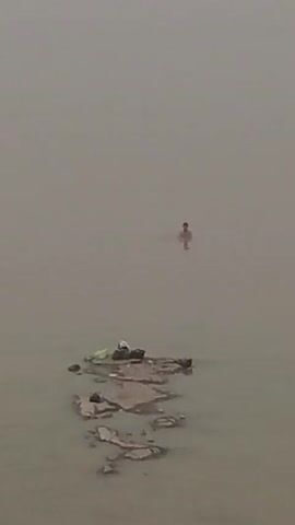 The video captures a hazy, overcast day with a body of murky water. In the distance, a person is submerged in the water, with only their head and shoulders visible. Closer to the foreground, a small, rocky island or sandbar protrudes from the water. On this island, several items are visible, including what appears to be a white plastic bag, a black object, and possibly some clothing items. The water is calm, and the overall impression is one of desolation and quietude.