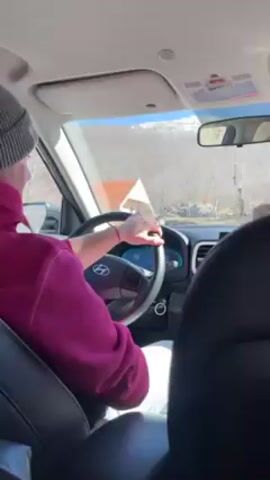 A person in a gray beanie and a dark pink hoodie is driving a Hyundai vehicle on a winding mountain road on a bright, sunny day. The dashboard is visible, showing a digital speedometer. Outside, the landscape features trees with bare branches and some snow-covered mountain peaks in the distance. The camera is positioned from the back seat, offering a view of the driver and the road ahead. Another person is seated in the passenger seat. The driver is focused on navigating the curves of the road.