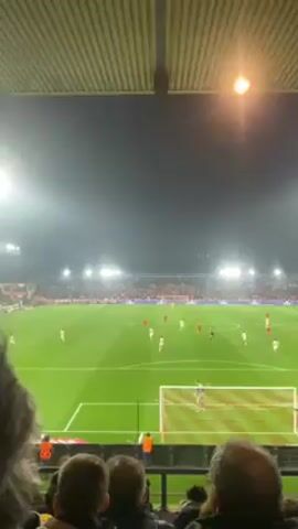 The video shows a soccer game at night, filmed from the stands. Players in red and white uniforms are visible on the green field. Bright stadium lights illuminate the field. Spectators are seated in front of the camera, partially obscuring the view. The stadium has a corrugated roof.
