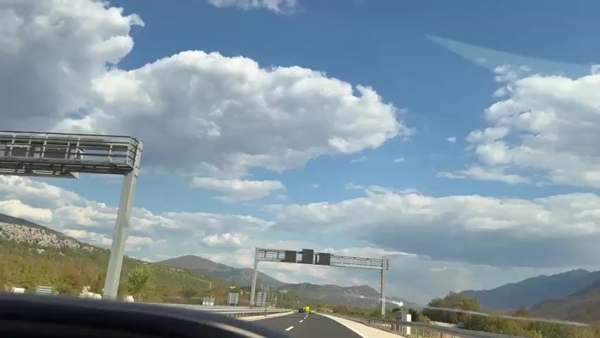 The video shows a view from a car driving on a highway. The sky is blue with large white clouds. Mountains are visible on both sides of the highway, covered with trees and vegetation. The road has multiple lanes, and there are guardrails along the side. An overhead gantry with traffic signs is visible. In the distance, there are some wisps of smoke or steam rising from the landscape. The car is moving at a moderate speed. The perspective is from the driver's seat, looking forward through the windshield.
