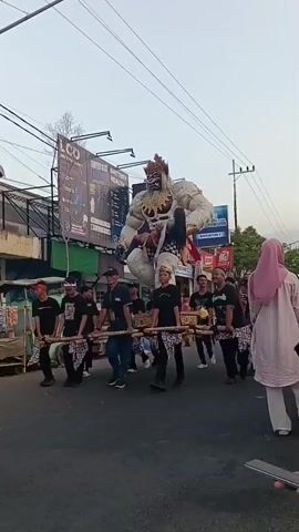 A group of people are carrying a large, elaborate effigy of a monstrous figure down a street. The effigy is white and appears to be made of fabric or paper-mâché, with a fearsome face, red and gold accents, and a long black-haired figure draped over its shoulder. The men carrying the effigy are wearing black shirts and patterned wraps around their waists. They are walking in a procession, some holding bamboo poles that support the effigy's platform. In the background, there are buildings, including a store with a red and white sign that reads "Alfamart." There are also power lines and streetlights visible. A few bystanders are also present, some watching the procession. The overall atmosphere suggests a cultural or religious event, possibly a parade or festival.