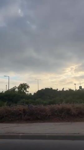 The video shows a view from inside a moving vehicle. The scene is dominated by a cloudy sky. In the mid-ground, there is a fence topped with barbed wire. Behind the fence, various buildings and structures are visible, some of which appear to be part of an airport or industrial complex. There are also streetlights and vegetation, including bushes and trees. The foreground shows a roadside with a pavement and a drain cover. The movement of the vehicle causes the scenery to shift and blur slightly, especially towards the edges of the frame. The lighting suggests it is either early morning or late evening.