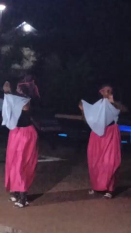 Two women dance outside at night. They are both wearing long pink skirts, black tops, and white pieces of fabric. They hold the fabric out and sway it as they move. There are plants and a few illuminated structures visible in the background. The scene is dark.