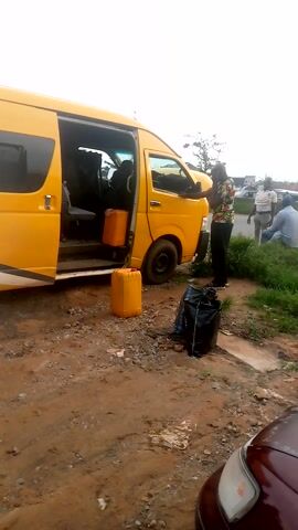 Here is a concise description of the video:

The video shows a yellow and white minibus parked on the side of a road. A man is standing next to the open side door, and there are two yellow containers and a black bag next to the van. A group of people are standing around the rear of the van, including a man wearing a blue uniform. The van has the logo "Anidis Ben Flo Ltd" on the rear. The ground is a mix of dirt, grass, and concrete. The sky is overcast. The video appears to be taken from a handheld device.