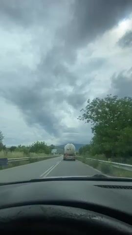 The image provides a first-person perspective from inside a vehicle, looking forward through the windshield at a two-lane asphalt road. The car's dashboard and a portion of the steering wheel are visible at the bottom of the frame.

The vehicle is following a white tank truck, which has multiple red and orange warning placards on its rear, indicating the transport of hazardous materials. On either side of the road, green trees and bushes line the view, separated from the roadway by metal guardrails. In the distance, hills or mountains are visible.

The sky is heavily overcast with dense, dark grey clouds, though some brighter patches suggest it is daytime.

Several signs are present:
*   On the left, a large blue and white billboard displays the word "СУХСТРОЙ" (SUKHSTROY) along with phone numbers and a website.
*   On the right, a red triangular warning sign with an exclamation mark indicates "other dangers."
*   Further down the road on the right, a blue directional sign with white arrows is visible, pointing towards different locations.

The scene depicts a drive through a green, somewhat rural landscape in Glavinitsa, Bulgaria, under cloudy weather conditions.