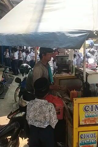 A person in a dark shirt and a black head covering is resting their hand on their head while looking up. They are seated next to a wooden cart. Behind them, several other people are visible, as are motorcycles and tents. The scene appears to be outdoors, possibly a market or event.