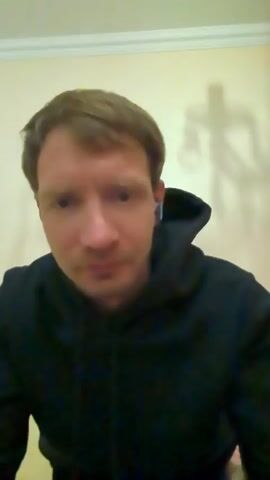 A man in a black hoodie is looking at the camera. He has short brown hair and appears to be wearing earbuds. A shadow of what looks like a robot or a person in a costume is visible on the wall behind him. The man makes a few facial expressions, including a neutral look, a smile, and a surprised expression. The background appears to be an indoor setting with a wall and a ceiling.