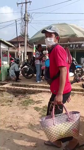 The video begins with a close-up of a person wearing a red shirt, black pants, a face mask, and a baseball cap, carrying a woven bag. The camera then pans to the right, showing a street scene with several parked motorcycles. In the background, there are small food stalls or shops with people gathered. One stall has a red umbrella and appears to be selling food. Further down the street, there is a building with a sign that reads "WARUNG MAKAN MBOKYO," indicating a food establishment. The overall environment appears to be an outdoor market or roadside area with a mix of commercial activity and people on foot and on motorcycles. The lighting suggests it is daytime.