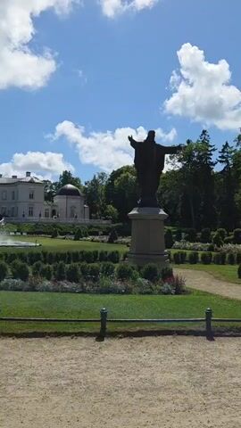 A video pans to the left, showing a large white manor house with a dome-shaped structure attached, set against a bright blue sky with fluffy white clouds. In front of the house, there is a formal garden with manicured hedges, flower beds, and a fountain. To the right of the house, a dark statue of a figure with outstretched arms stands on a pedestal. The foreground is a gravel path with a low green fence.
