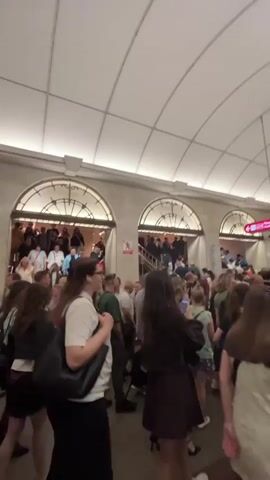 The video shows a crowded indoor scene, likely a subway station. A large group of people are visible, many of them ascending a wide staircase through arched doorways. The architecture features a curved ceiling with recessed lighting. People are dressed in casual clothing. In the foreground, some individuals are walking towards the camera. The overall impression is one of a busy thoroughfare with a significant number of commuters or travelers.