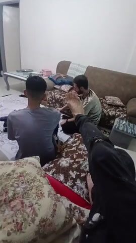 The video shows three men in a room. One man in the foreground, partially obscured by a pillow with a floral pattern, has his right hand extended towards the camera, palm facing forward. He is wearing a dark long-sleeved shirt. Behind him, another man sits facing away from the camera, wearing a gray t-shirt. He is looking towards a third man who is seated on a sofa, facing the man in the gray t-shirt. This third man has a beard and is wearing a patterned shirt. He appears to be holding a phone or similar object. The room has a sofa with patterned cushions, a coffee table with items on it, and a wall with a light switch. The lighting is dim. The man in the gray t-shirt turns his head to look directly at the camera.