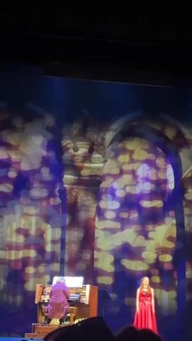The media file shows a live performance on a stage. A person is playing a pipe organ, and a young woman in a red dress is singing. The background is a projection of a grand architectural space, possibly a cathedral, with arches and intricate details. The projection is illuminated with colorful, bokeh-like lights in shades of purple, yellow, and gold, creating a dreamlike atmosphere. The lighting on stage is dim, with spotlights focusing on the performers. The audience is partially visible in the foreground, silhouetted against the stage lights. The overall mood appears to be one of artistic expression and a formal musical performance. The text "00:00" is visible in the top left corner, indicating a timestamp. Given the location context of Odesa, Ukraine, this could be a performance in a concert hall, church, or theater within the city. The time of day is not discernible, but the indoor setting suggests it could be any time. The emotions conveyed are likely a mix of solemnity and beauty, appropriate for an organ and vocal performance.