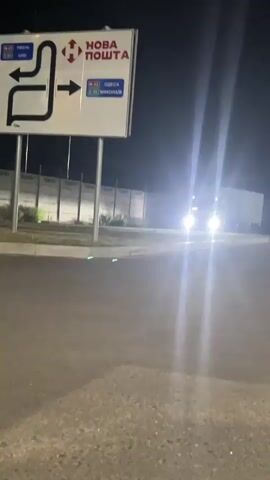 A white truck approaches a barrier arm that is lowered. The truck has its headlights on, illuminating the area. There is a road sign in the background with directional arrows and text that appears to be in Ukrainian. The sign indicates directions to "Nova Poshta", "Kyiv", "Odesa", and "Mykolaiv". The truck is in motion and appears to be coming to a stop at the barrier. The environment is dark, suggesting it is nighttime.