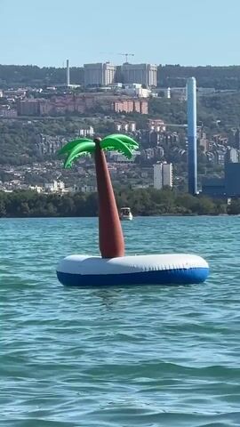 An inflatable island with a palm tree floats on the water. The island is white with a blue border. The palm tree is brown with green leaves. In the background, there is a city skyline with buildings and trees. The water is blue and choppy. A small boat is visible in the distance.