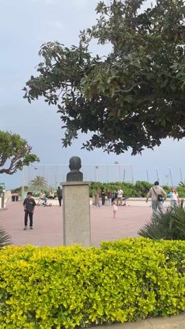 The video shows a public square.  A bust sits atop a pedestal surrounded by a low hedge. The square has a pattern of red brick. People stroll, sit, or engage in recreational activities such as playing with a scooter. A boy dribbles a basketball.  Another boy watches with hands on his hips. Beyond the square is a fence, some shrubbery, and the sea. The sky is overcast. Several trees are visible in the scene.