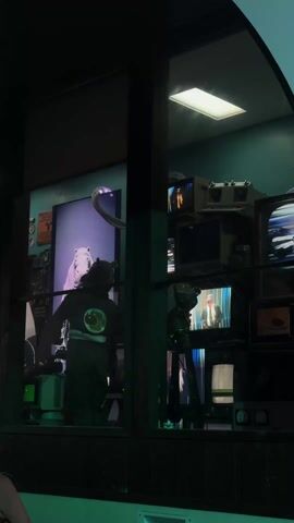 The video shows an exterior view of a building window filled with various television sets, broadcasting different images. A figure wearing a dark jacket with a circular symbol on the back stands in front of a display featuring a person in a pig mask. The camera then pans upward to reveal the building's exterior, including a sign with indistinct text and a round window. The video concludes with an exterior shot of an outdoor event, featuring palm trees, lighting rigs, and what appears to be a crowd of people against a dusk sky.