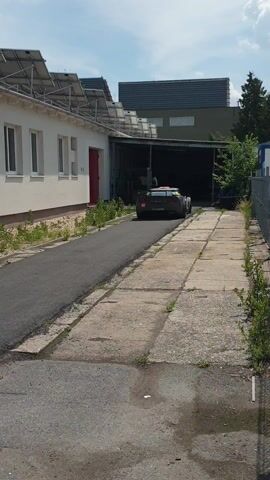 A black sports car is parked in front of a garage, with its rear facing the camera. The car has red taillights and a spoiler. There are solar panels on the roof of a building to the left of the garage. The ground is paved with concrete and asphalt. The video is short and static, with no discernible action or interaction.