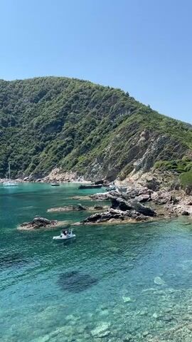 A drone shot pans across a clear, turquoise bay dotted with boats. Rocky outcrops jut out of the water, and a lush, green mountain slopes down to the coastline. In the foreground, a small inflatable boat with two people aboard bobs gently on the waves. Sailboats are anchored in the distance. The sky is a bright, cloudless blue.