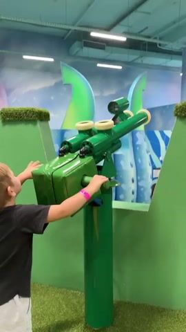 A young boy wearing a black t-shirt and white shorts looks through a green toy telescope in an indoor playground. The playground features bright blue and green decorations, including large tentacle-like structures and faux grass. The boy's left hand is on the toy telescope's handle. The camera is positioned behind and to the right of the boy. The video is a vertical screen recording.
