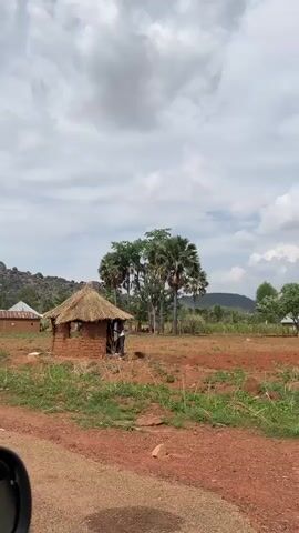 The video shows a rural landscape with a road in the foreground. On either side of the road are dirt fields with scattered vegetation, small trees, and some buildings. The buildings appear to be simple, possibly made of brick or mud, with some having tin roofs. In the background, there is a rocky outcrop under a partly cloudy sky. As the video progresses, a woman dressed in colorful clothing walks into view from the side of the road. She gestures with her arm as the camera passes by. The environment seems to be a village or rural setting, with signs of agricultural activity.