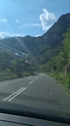 The video shows a view from inside a car driving on a paved road through a mountainous, forested area. The sun is shining brightly, with rays of light visible and lens flares on the camera. The road curves to the right, and the mountainside is covered in green trees and vegetation. The sky is blue with scattered white clouds. The dashboard of the car is visible at the bottom of the frame.