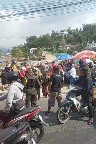 The video shows a crowded outdoor scene, possibly a street market or a gathering. There are many people, some wearing traditional clothing like hijabs. Several motorcycles are visible, some with riders. In the background, there are buildings and hills covered with vegetation, suggesting a rural or semi-rural environment. The sky is cloudy. Some people appear to be standing and talking, while others are on motorcycles. There are also some market stalls or tents set up. The overall impression is of a lively, populated area.