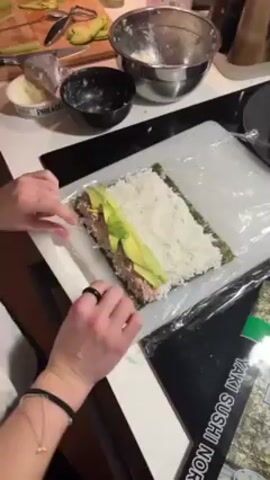 The video shows a person making sushi on a white countertop. The person, wearing a watch and bracelets, is rolling a sushi roll on a clear plastic sheet covering a nori sheet spread with rice and topped with avocado slices and tuna. Bowls with ingredients like cream cheese and fillings are visible along with a package of Yaki Sushi Nori. Other utensils and avocado slices are present on a cutting board nearby.