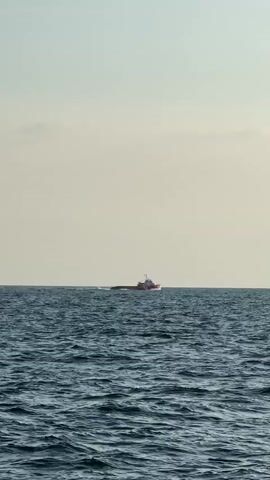 A medium shot captures a red and white boat traveling across a vast expanse of blue, choppy water under a pale, overcast sky. The boat is positioned in the center of the frame, moving from left to right. The horizon line is clearly defined, separating the water from the sky. The boat is the only object in the water, and no people are visible on its deck. The scene suggests a tranquil, open-water environment with mild weather conditions.