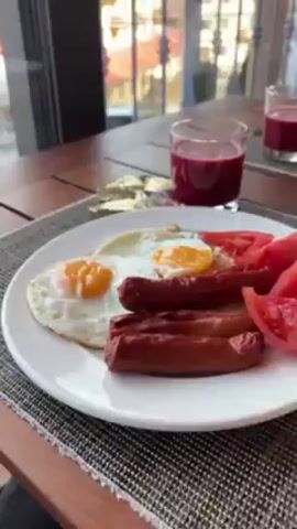 The video shows a breakfast scene next to a window. On a brown wooden table, there's a white plate with two sunny-side-up eggs and several sausages. Next to it, there are butter packets and a glass filled with red juice. The camera then pans towards the window showing a view of buildings, a parking lot with cars, a flag with red and green stripes, a town, and distant snow-capped mountains under a partly cloudy sky.