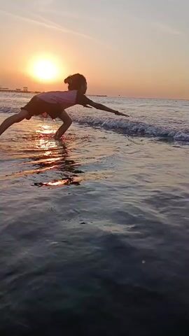 The media file depicts a young girl playing in the shallow ocean water at sunset. The sun, a bright orb, is visible on the horizon, casting a warm, orange glow across the sky and reflecting on the water's surface. Gentle waves lap at the shore, creating a serene and peaceful atmosphere. The girl is silhouetted against the vibrant sunset, her playful movements creating splashes in the water. She appears to be enjoying herself, her posture suggesting an active engagement with the sea. The scene is set on a beach, with a distant pier or structure visible on the left side of the frame. The overall mood is tranquil and idyllic, capturing a moment of childhood joy amidst a beautiful natural setting. There is no visible text in the media file.