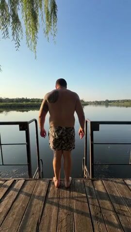 A man in swim trunks stands on a wooden pier facing a body of water. He stretches his arms out to the sides and then jumps into the water, creating a splash. A black and white dog watches from the pier. The scene is set on a clear day with a blue sky and trees in the background.