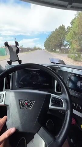 The video provides an interior view from the driver's seat of a large truck. The driver's left hand is on the steering wheel, which displays the Western Star logo. The dashboard features a digital speedometer showing speeds between 24 and 31 mph. A black phone mount is attached to the windshield, and a phone is mounted in it. The truck is driving down a paved road lined with trees and residential fences. The sky is cloudy with patches of blue. The video appears to be a first-person perspective of driving the truck through a suburban or semi-rural area.