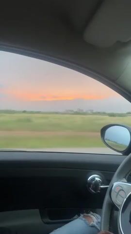 The video captures a view from inside a car driving through a rural landscape near Sidi Abdallah Ghiat, Morocco, during sunset. The sky is painted with soft orange, pink, and light blue hues visible through the car's window. Below the horizon, green fields stretch into the distance, with blurred structures suggesting distant buildings. The car is in motion, evidenced by the horizontal blur of the outside scenery.

Inside the vehicle, parts of the dark dashboard, a chrome door handle, and a grey steering wheel with controls are visible. A person's leg, clad in distressed denim jeans, can be seen near the steering wheel. A hand enters the frame, holding a green and yellow apple, and then tosses it out of the open window. The car's side mirror reflects the moving landscape and the vibrant sky. The scene depicts a casual moment during a drive as daylight fades. No visible text is present.
