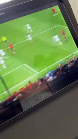 This media file captures a snippet of a live football (soccer) match being viewed on a tablet. The tablet screen displays a green football field with several players in red and white uniforms. The game appears to be intense, with players actively engaged in the match. In the foreground of the tablet screen, a crowd of spectators can be seen, many with their arms raised in excitement, suggesting a passionate atmosphere.

The scene is set indoors, as indicated by the tablet being held and the reflection on its screen. The tablet itself is housed in a dark protective case. The visible text on the screen includes "EN VIVO" (Spanish for "LIVE"), and "T" within a red circle, which is the logo for Televisa Deportes, a sports broadcasting channel. Additionally, there are sponsor logos visible on the stadium advertising boards, including "FIFA", "America's B", and "Network". The time of day cannot be determined from the visual information alone. The overall emotion conveyed is one of excitement and engagement with the sporting event. The location context of Almada, Portugal is provided externally.