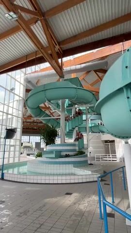 This video shows the interior of an indoor water park or swimming pool. The central feature is a large, light blue/turquoise water slide complex, featuring a prominent spiral slide. A white spiral staircase with blue and white tiled accents leads up to the slide platforms, where several people, mostly children, are visible queuing to use the slides. Another part of a slide, also light blue, is visible to the right.

At the base of the slide structure, there's a shallow pool with blue and white tiled edges, and some green plants are arranged around its perimeter. The building features a high ceiling with exposed wooden beams and trusses, suggesting modern architecture. Large glass windows on the left side provide ample natural light, indicating it's daytime. The floor throughout the area is tiled in white and light blue. People are gathered around the base and on the stairs, suggesting a busy recreational environment. No visible text or specific branding is discernible.