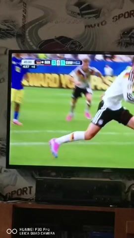 This video shows a close-up, somewhat blurry, shot of a television screen displaying a soccer match. The score is visible at the top left of the screen: GER 3, GUA 1. The time displayed is 45:00 +4. Several soccer players are on the field, wearing white jerseys with black stripes on the sleeves and black shorts. Their socks are white with black stripes, and they are wearing bright pink soccer cleats. One player in the foreground is kicking a soccer ball. The background shows a blurred crowd and a green soccer field. The television is placed on a stand, below which are some electronic devices. The wallpaper in the background behind the television has a repeating pattern of cars and the words "AUTO SPORT."