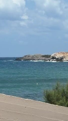 A view of the sea and a rocky island. The water is a deep blue-green and there are some waves breaking on the shore of the island. The island is covered in dry, reddish-brown vegetation and some larger grey rocks. The sky is blue with some white clouds. In the foreground, the edge of a tan-colored structure is visible, with some green bushes in front of it. A small red buoy is visible in the water near the island.