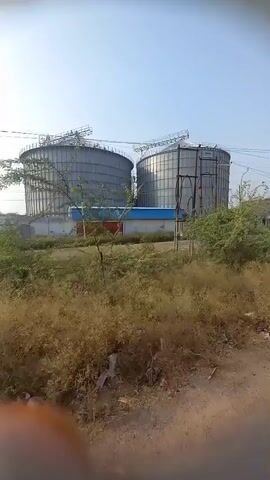 The video captures a view of two large, cylindrical, metal silos, likely grain storage facilities, set against a clear, blue sky. The silos are situated behind a low, blue and white building, and are surrounded by dry, grassy vegetation. Utility poles are visible along the roadside, with wires running between them. The camera appears to be moving, potentially from a vehicle, as the scene gradually shifts. The overall environment suggests a rural or industrial area.