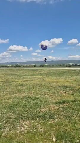 The video shows a wide shot of a grassy field with a blue and white parachute and two individuals on the ground. The sky is blue with scattered white clouds. The parachute is partially visible, with its lower section being blue and the upper section being white. Below the parachute, a person appears to be landing. To the left of the parachute and closer to the left edge of the frame, another person is sitting or kneeling on the ground. The field is a mix of green and patches of brown earth. In the distance, there's a strip of land with some trees and a low, distant hill. The scene suggests a skydiving or paragliding activity taking place in an open, rural environment.