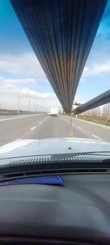 From the interior of a vehicle traveling on a paved multi-lane highway, the driver is following a flatbed truck carrying a large load of rebar. The rebar is bundled and stacked vertically on the front of the truck, partially obstructing the driver's view. There are streetlights lining the side of the highway and other vehicles are visible further ahead. The sky is blue with some clouds.