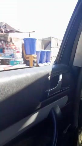 This is a first-person perspective video from inside a vehicle, looking out the passenger-side window. The scene appears to be an outdoor, dusty area with a bright, clear blue sky, suggesting it is daytime and fair weather.

The immediate foreground is dominated by the interior of the car door, including the armrest, door handle, and window controls. Through the window, we see a scene that suggests a temporary settlement or encampment. Structures covered with white tarpaulin, possibly tents or makeshift shelters, are visible. Some of these structures have the UNICEF logo printed on them, indicating humanitarian aid or a refugee camp.

Several people and objects are observed moving through the scene. In the early frames, a silver station wagon drives by. Later, a man with a beard wearing a cap and a vest rides a bicycle. Another person in a red cap and a blue shirt walks past. In the background, someone is sitting under a blue tarp structure.

The overall impression is one of a busy, temporary living environment, possibly in a region experiencing displacement or in a humanitarian context. The presence of the UNICEF branding is a significant detail. The activity appears to be everyday life within this settlement.