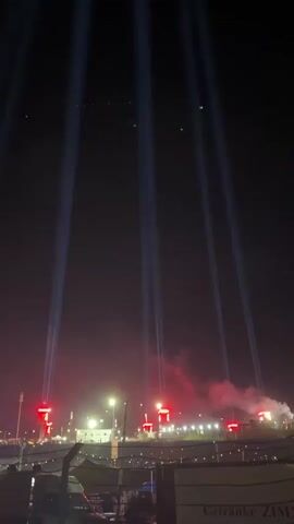 A nighttime scene unfolds with several powerful white spotlights cutting through the dark sky from a low vantage point, their beams diverging upwards. Below the beams, a line of red lights are visible, positioned along a structure that appears to be a stadium or event venue. Red smoke or haze drifts around these red lights. In the foreground, out of focus, are elements of a barrier and some signage, one piece of which appears to have German text. The overall atmosphere suggests an outdoor event or concert taking place at night.