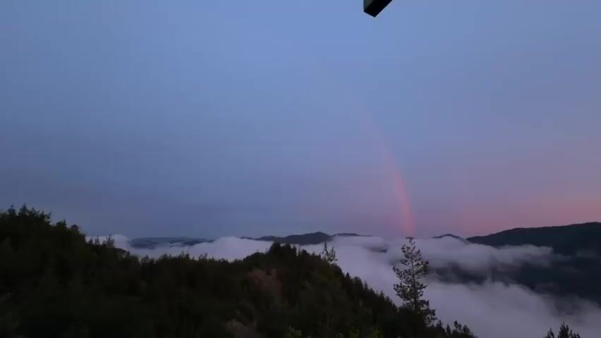 This is a landscape video capturing a view of a valley at dusk. The sky is a gradient of deep blue at the top transitioning to pale pink and orange near the horizon. A faint rainbow arcs across the sky, with the red part most visible. Below the sky, a layer of thick white clouds blankets the valley, obscuring the lower parts of the mountains. The tops of dark green, forested mountains emerge from the clouds. In the foreground, dark green trees and foliage are visible. The video is taken from a high vantage point, possibly a balcony or overlook, as a dark rectangular object is partially visible in the top center of the frame, suggesting a structure or fixture. There are no people or animals visible in the scene.
